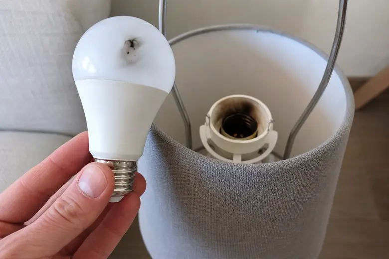 Main tenant une ampoule LED grillée devant une lampe de salon, avec le culot noirci visible