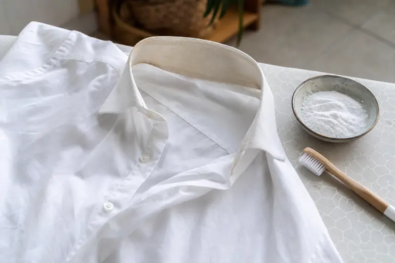 Chemise blanche posée à plat avec un col légèrement jauni, à côté d'une brosse à dents et de bicarbonate de soude
