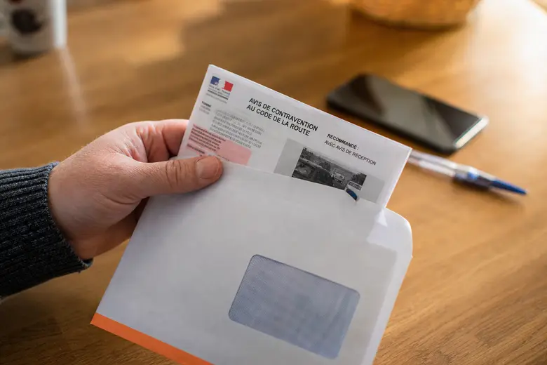 Main tenant un avis de contravention routière à côté d’une enveloppe recommandée, sur une table en bois