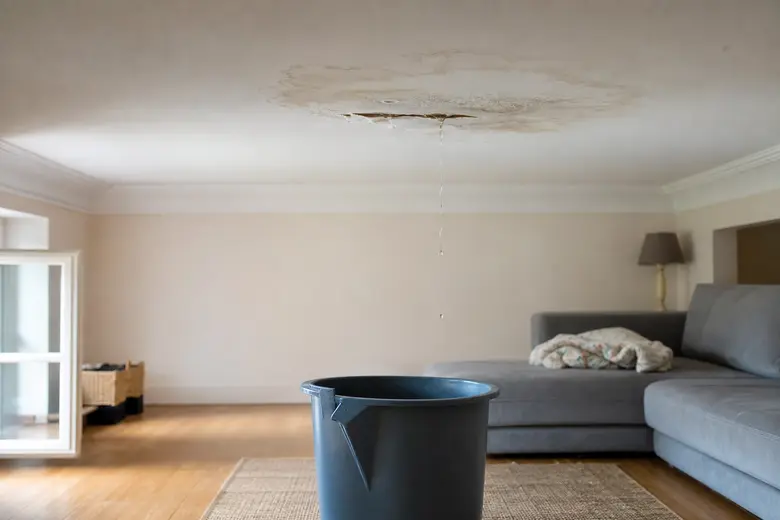 Tache d'humidité et infiltration d'eau sur un plafond blanc dans un salon, avec un seau posé au sol