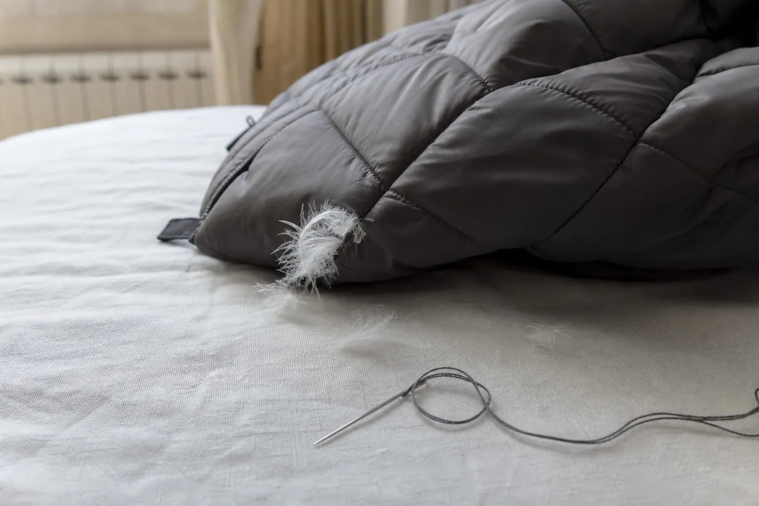 Doudoune posée sur un lit avec quelques plumes qui s'échappent d'une couture, à côté d'une aiguille et de fil