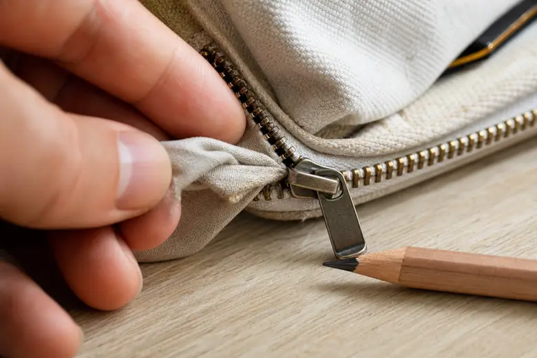 Main dégageant délicatement un tissu coincé dans une fermeture éclair, avec un crayon à papier posé à côté