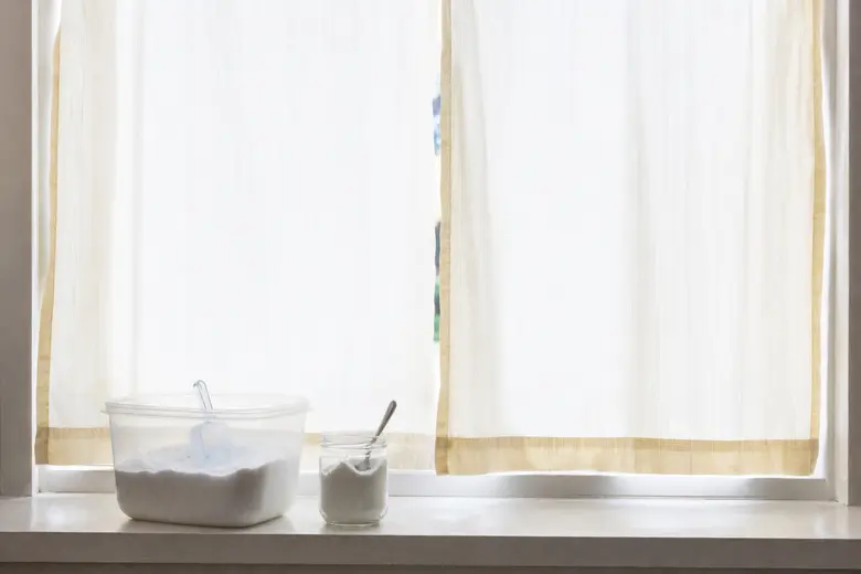 Rideaux blancs légèrement jaunis pendus devant une fenêtre lumineuse, avec du bicarbonate de soude posé sur le rebord