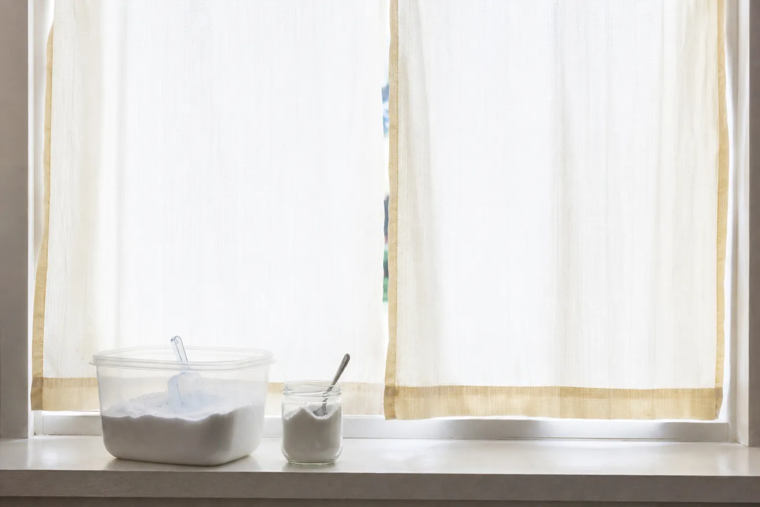 Rideaux blancs légèrement jaunis pendus devant une fenêtre lumineuse, avec du bicarbonate de soude posé sur le rebord
