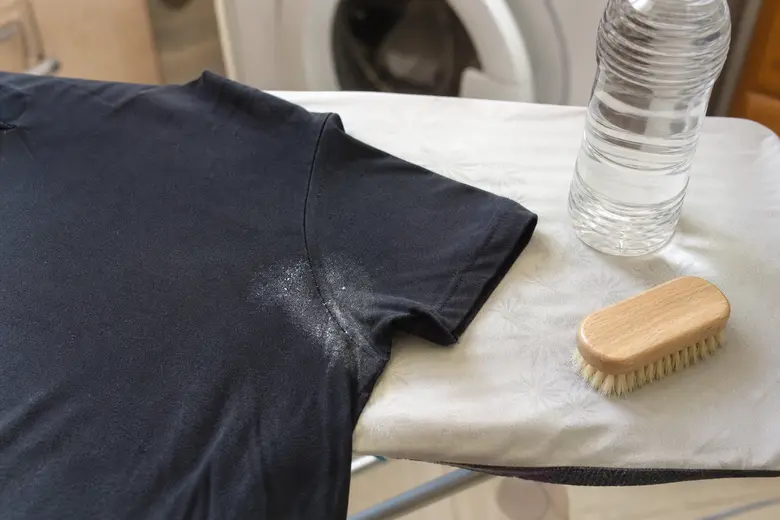 Tee-shirt noir avec une trace de déodorant sous l’aisselle, posé sur une planche à repasser à côté de vinaigre blanc et d’une brosse à vêtements